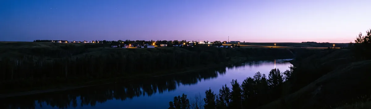 Generated picture: blue hour over a prairie river valley, a dark river below and a few lights of a small town on the far rim.